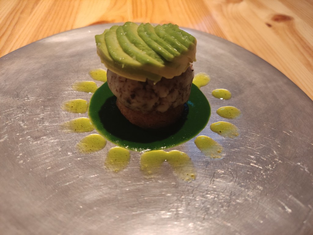 A silver plate with a pool of green sauce, a small biscuit, a dollop of potato, and sliced avocado on top