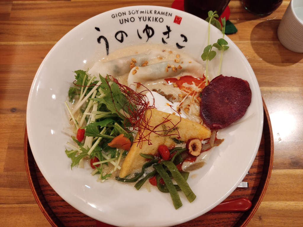 A creamy broth topped with two gyoza, a beetroot crisp, a triangle of tofu, spicy oil, and lots of vegetables