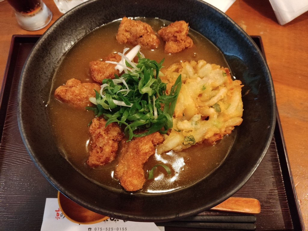 A bowl of brown broth topped with a large pieces of tempura, lots of green spring onion, and several crispy looking battered nuggets