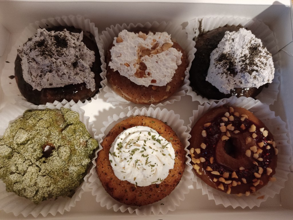 Six donuts in a box, the middle one on the bottom row is topped with a spiral of cream cheese and green herbs