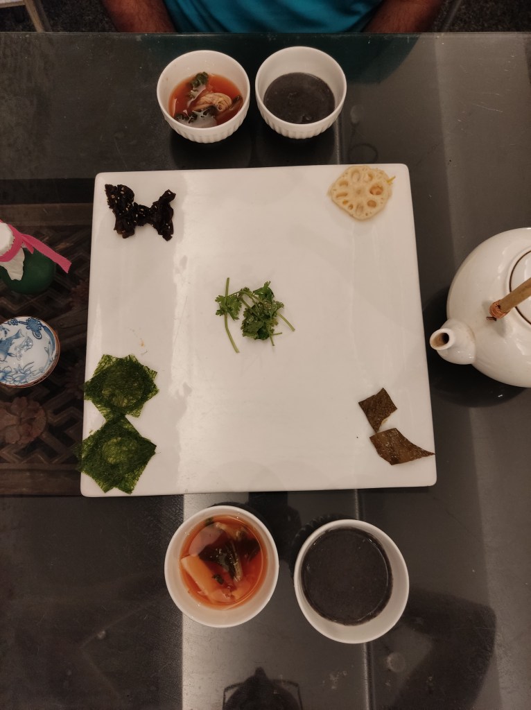 A large square white plate with a few pieces of food in each corner and some coriander in the middle. There are also two small bowls of soup at opposite sides of the table.