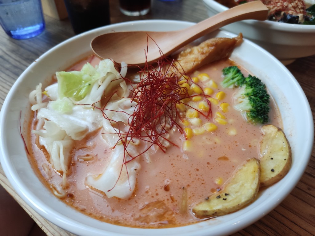 A bowl of pale red broth topped with sweetcorn, potato, broccoli, tofu, cabbage, and chilli threads
