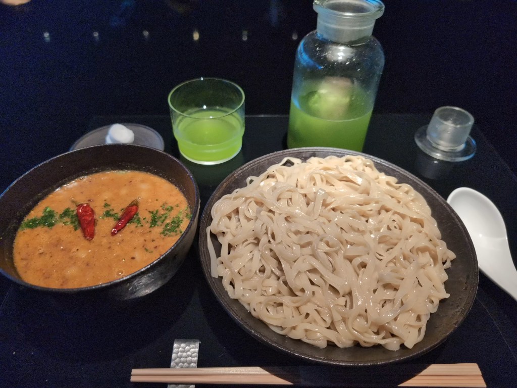 A large bowl of wide flat noodles and a separate bowl of orange broth topped with chillies. There's a bright green tea in the background.