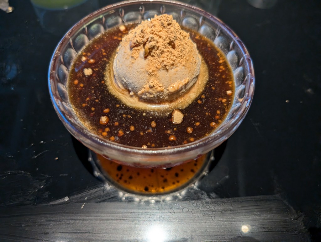 A scoop of beige ice cream topped with a powder the same colour and sitting in a darker brown sauce