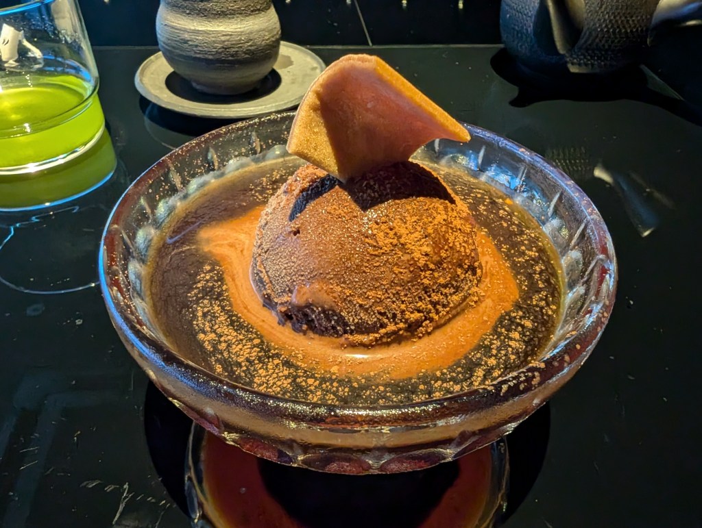 A scoop of dark brown ice cream dusted with a beige powder and topped with a wafer and sitting in a dark brown sauce