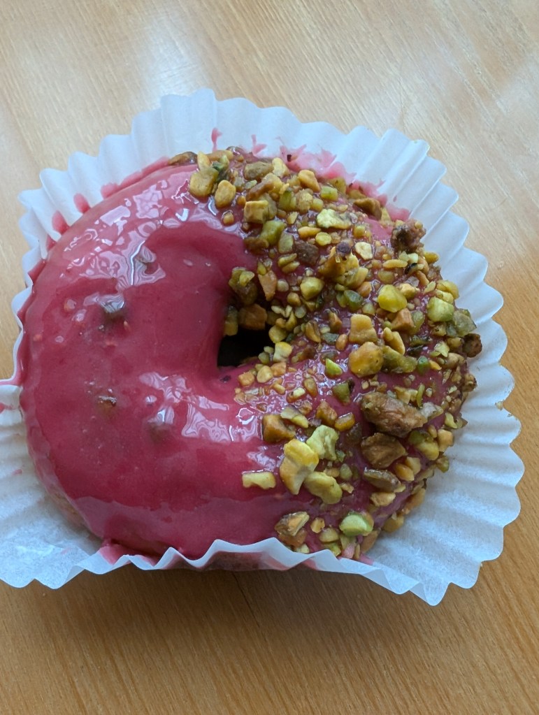 A donut with a sticky bright pink icing and one half covered with chopped pistachios