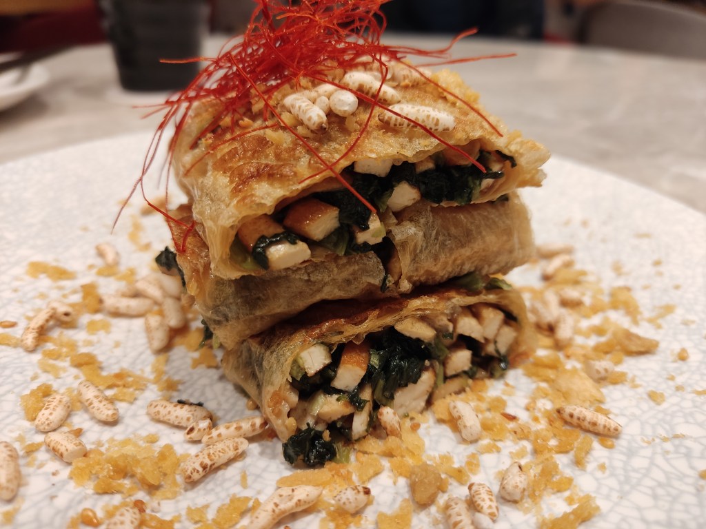 A stack of three bean curd skin rolls stuffed with greens, tofu, and puffed rice
