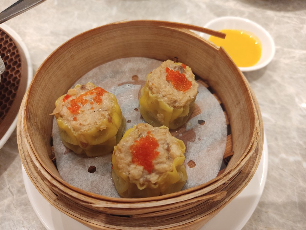 Three dumplings in a steamer, yellow wrappers with a beige filling, open at the top and topped with tiny orange balls