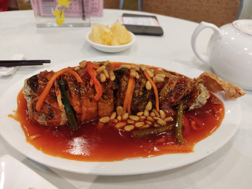 A large vaguely fish shaped dish with a crispy skin sitting in a bright red sauce and topped with pine nuts and a few shredded vegetables