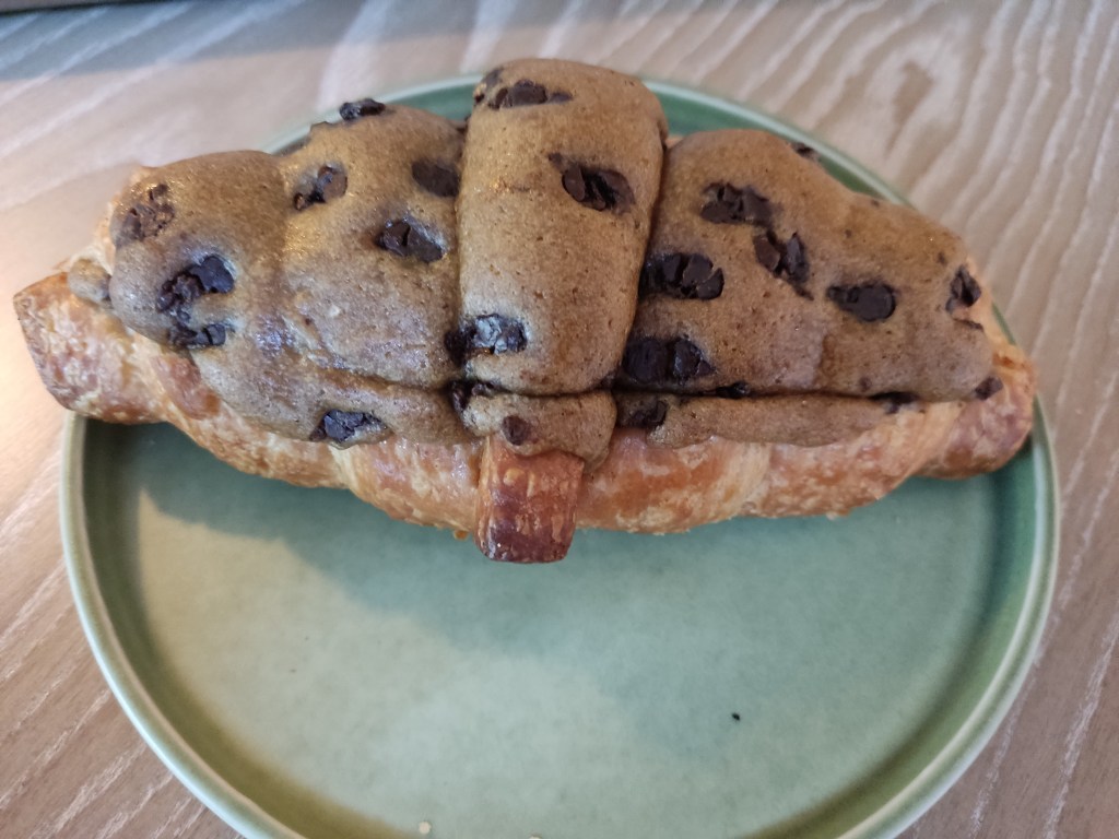 A crookie that looks like a croissant, topped with a hard looking dark light brown shell studded with chocolate chips