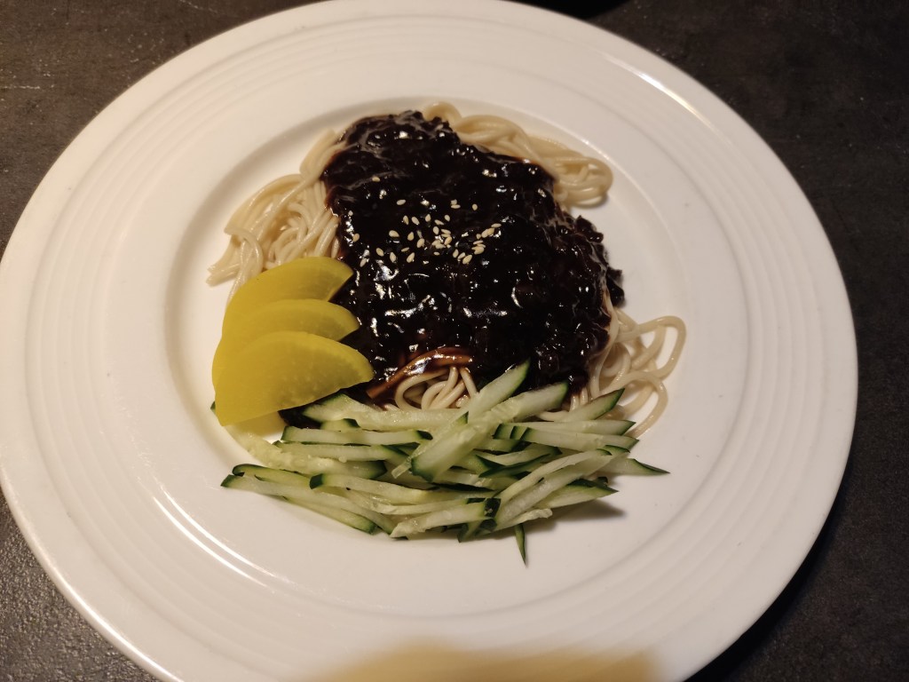 A plate of noodles topped with a dark brown sauce alongside sliced cucumber and pickles