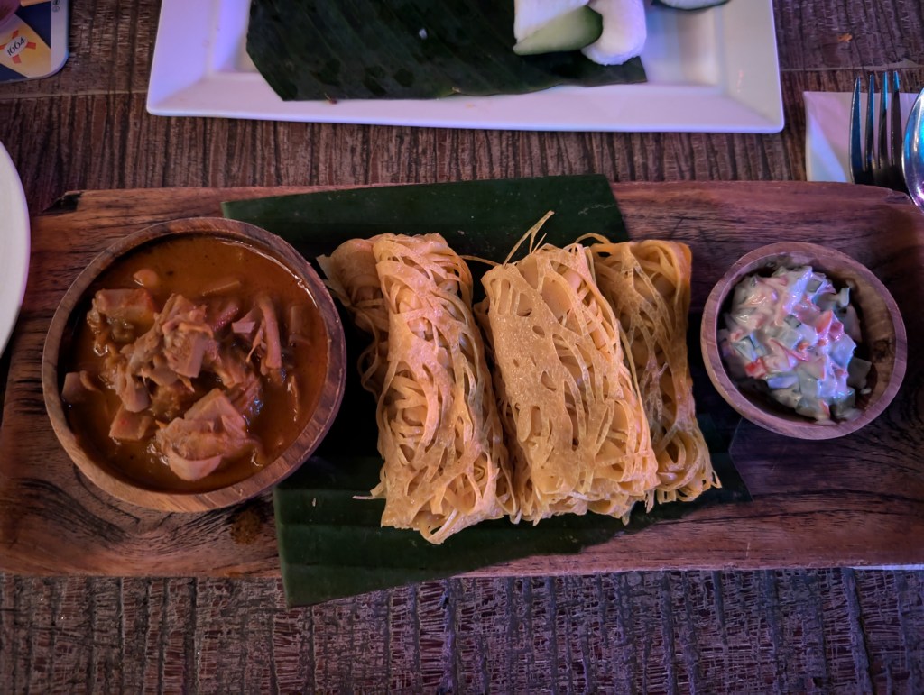 A pot of jackfruit curry, a pot of raita, and three lacy looking breads