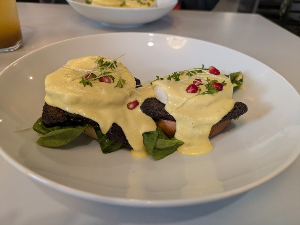 Two toasted English muffins topped with tofu bacon, tofu egg, spinach, and yellow hollandaise sauce