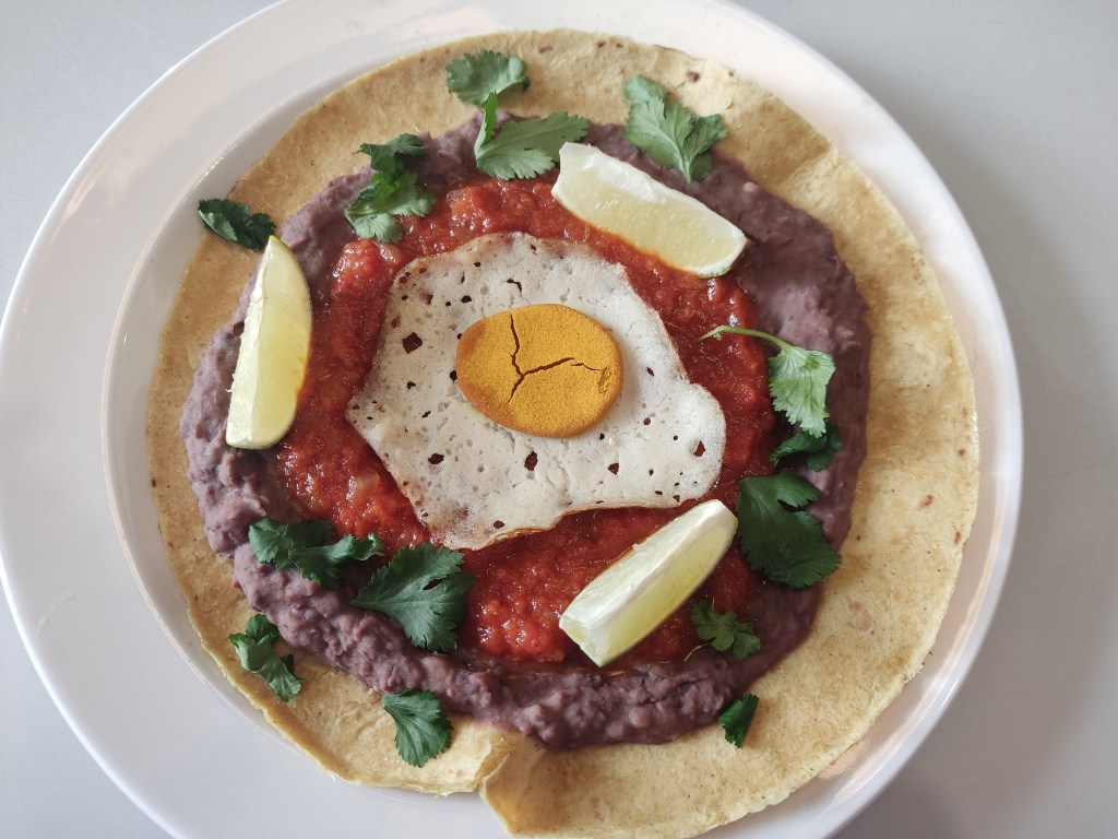 A tortilla topped with refried beans, tomato sauce, a mock fried egg and lime wedges