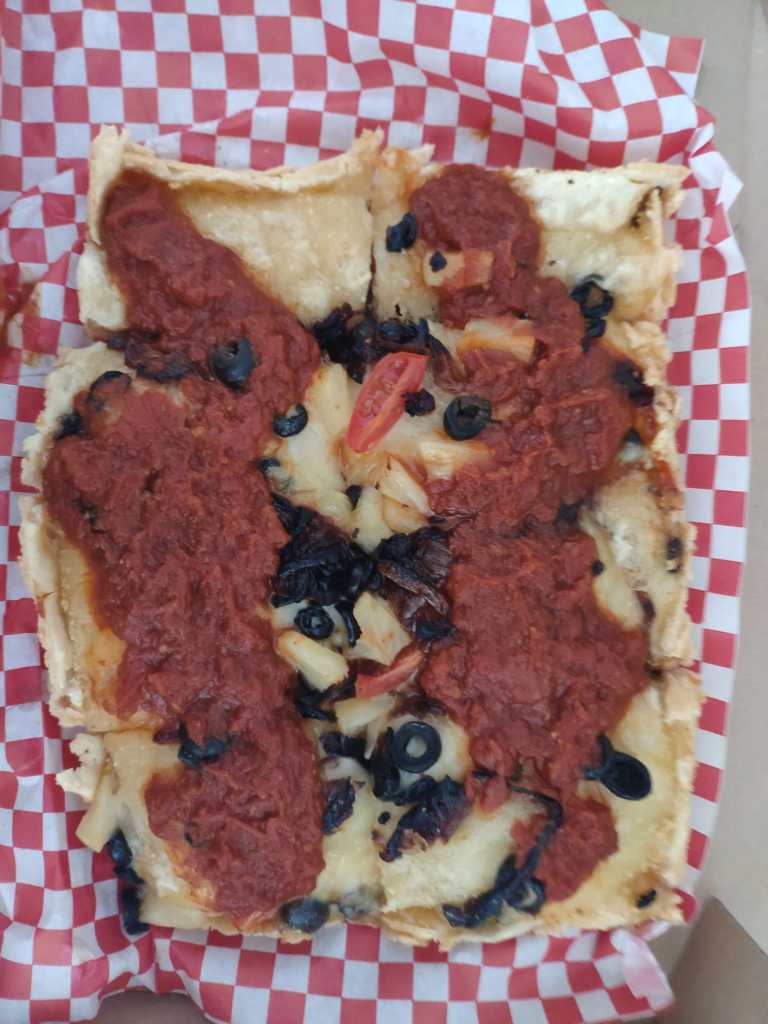 A square pizza cut into eighths covered in vegan cheese and served with tomato sauce scooped on and topped with pineapple, black olives, onions, and cherry tomatoes
