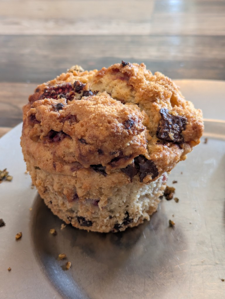 A muffin with a fluffy base and crisp, craggy top, with chunks of dark chocolate and pieces of raspberry visible