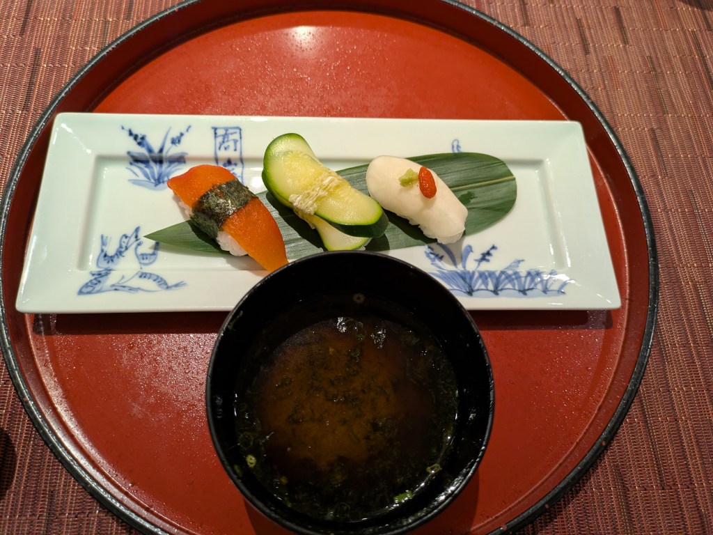 Three pieces of sushi each topped with a slice of a different vegetable - red pepper, courgette, and turnip. Also a small bowl of broth.