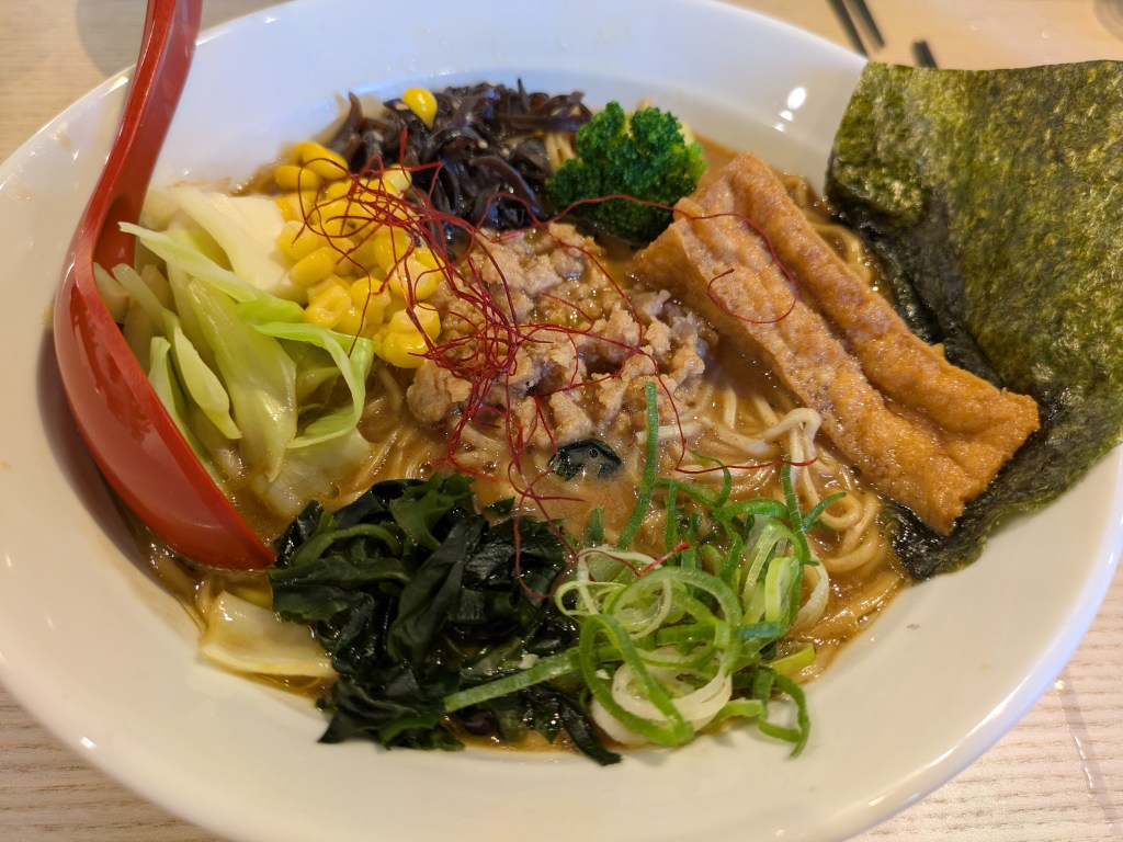 A bowl of ramen with a brown broth topped with seaweed, tofu, and lots of different vegetables