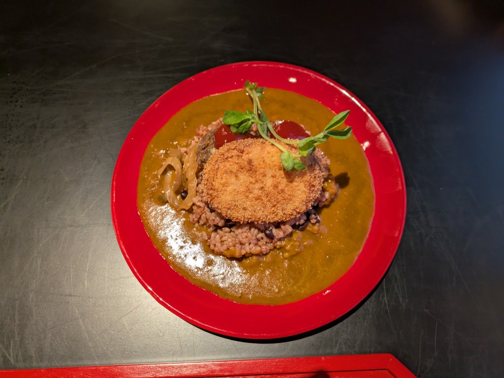 A big plate of curry sauce with a mound of rice, some meatballs, and a croquette on top