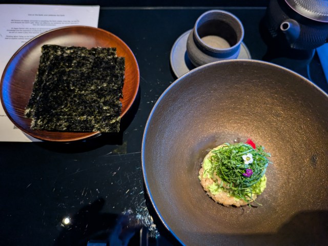 A piece of nori on a side plate and a bowl containing a small mound of rice topped with vegetables