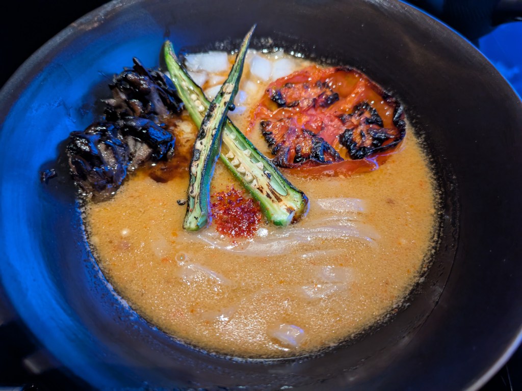 A big bowl or ramen with a yellow coloured broth. It's topped with mushrooms, okra, and a large piece of grilled tomato.