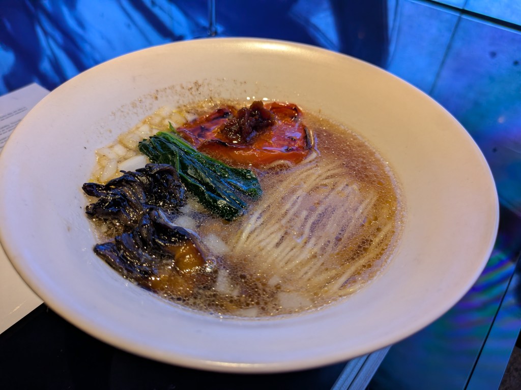 A big bowl of ramen topped with mushrooms, green leafy vegetables, and a large piece of tomato. A big portion of noodles is visible under the broth