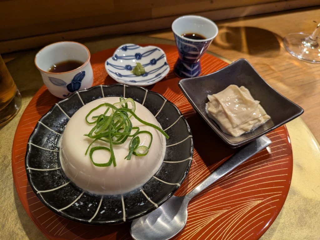 A tray containing several dishes. There is a large serving of silken tofu topped with spring onions, with a brown dressing on the side. There's also a dish with a tiny helping of wasabi. Another dish contains pieces of yuba, served alongside a little cup with brown dressing