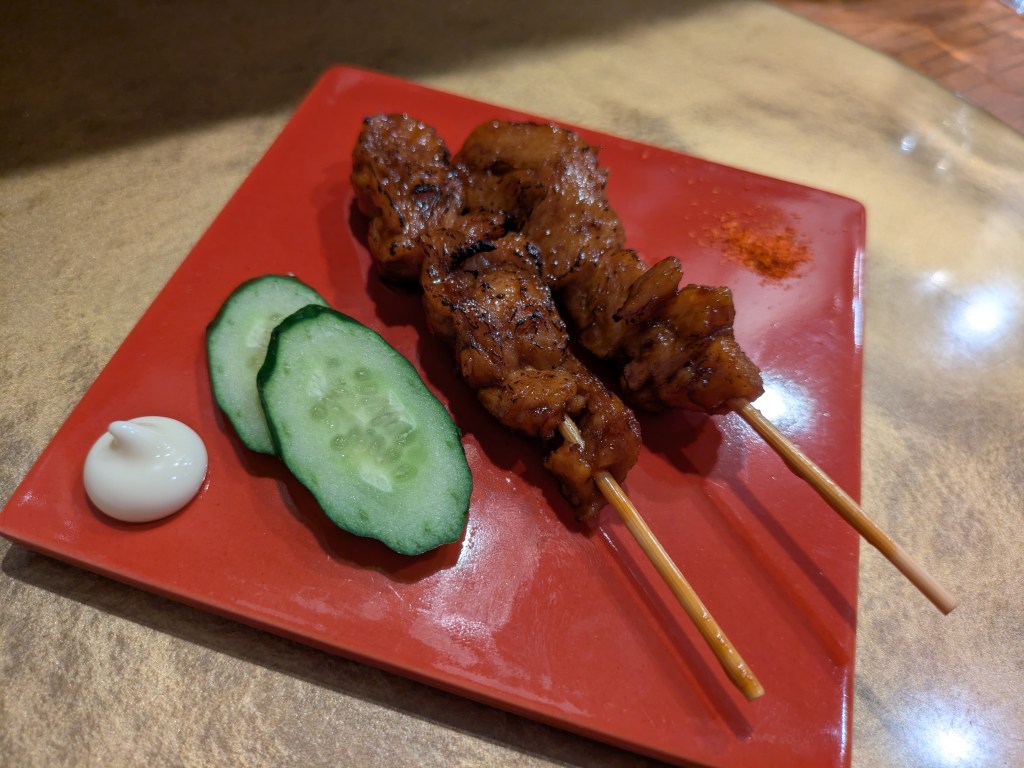 A plate with two meaty skewers, two pieces of cucumber, and a dollop of white sauce