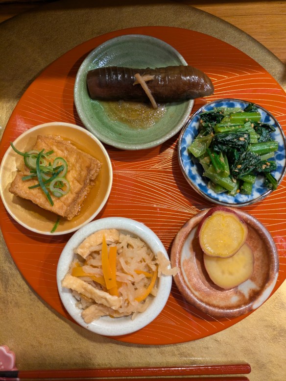 A tray with five small plates. One is a large piece of aubergine, one has lots of small pieces of greens, one has two slices of sweet potato, one has some pickled radish and tofu, and one has a big piece of fried tofu.