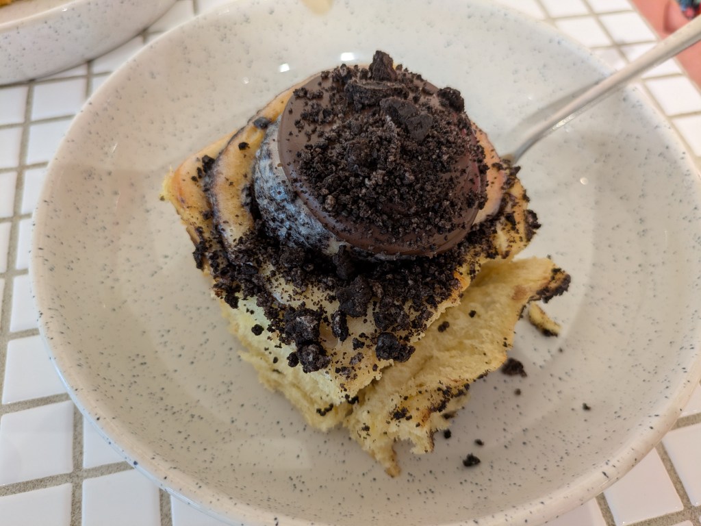 Cinnamon roll topped with a big dollop of chocolate cream and crumbled Oreos