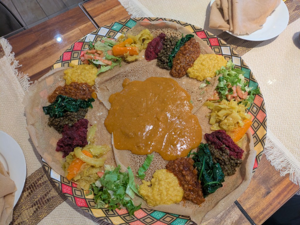 Three large injera overlapping like a Venn diagram, topped with dollops of numerous different lentil and vegetable dishes, with a big pile of sauce in the middle