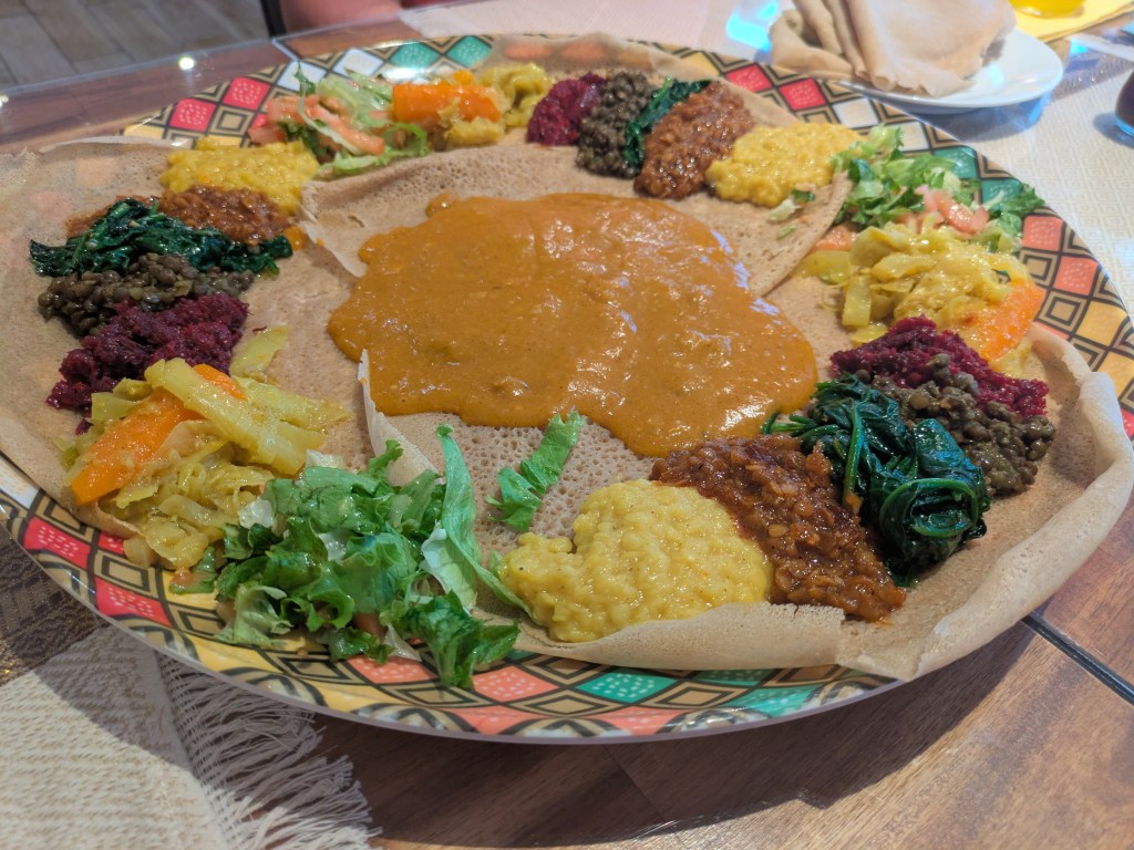 Three large injera overlapping like a Venn diagram, topped with dollops of numerous different lentil and vegetable dishes, with a big pile of sauce in the middle