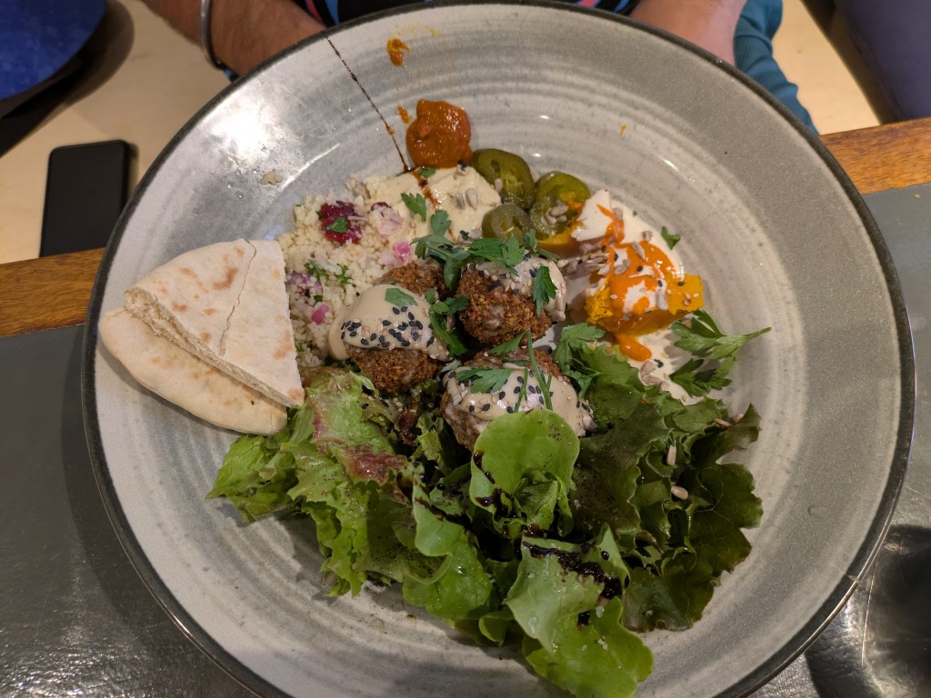 A bowl with salad, two shards of pita, falafel, couscous and salad