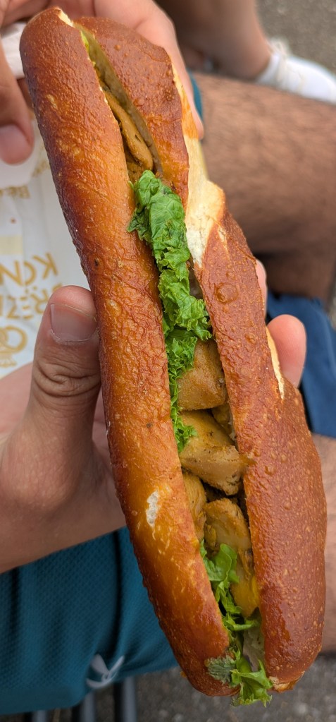 A long pretzel bun filled with salad leaves, mock meat, and a bright yellow curry sauce 