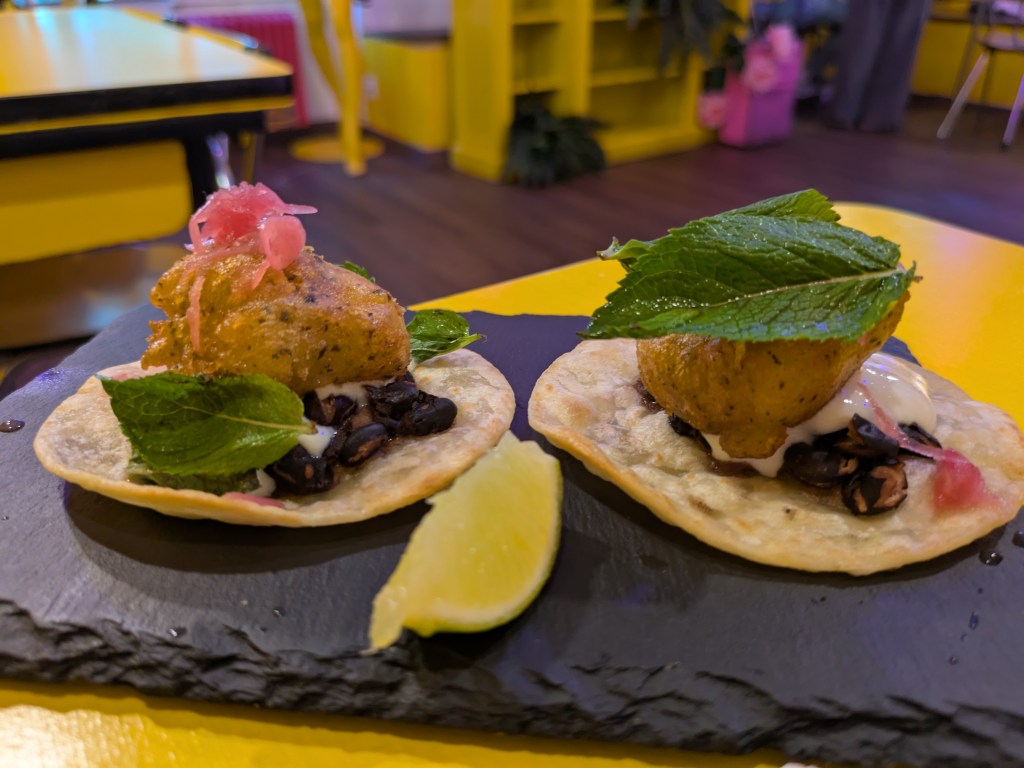 Two tacos topped with black beans, beer battered tofu, vegan labenh, and garnishes