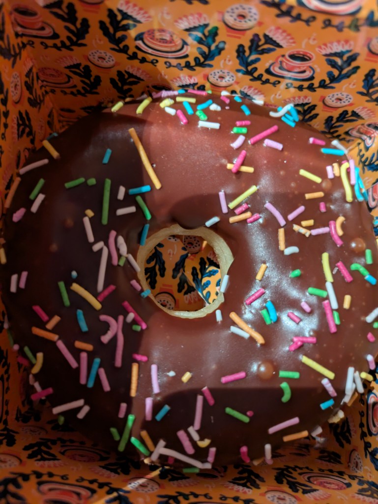 A takeaway box containing a ring doughnut covered in chocolate and sprinkles