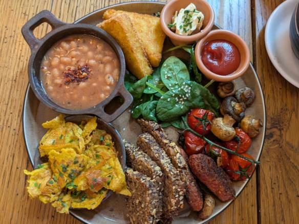 A big plate with hash browns, baked beans, tofu omelette, sausages, bread, spinach, tomatoes, mushrooms, butter, and sauce