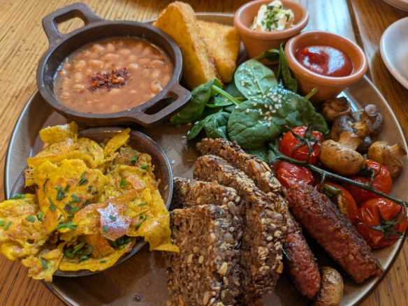 A big plate with hash browns, baked beans, tofu omelette, sausages, bread, spinach, tomatoes, mushrooms, butter, and sauce