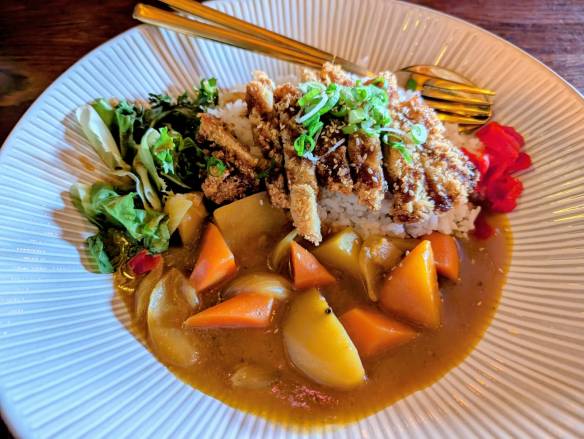 A bowl of curry with rich sauce, large chunks of root vegetables, rice, salad, and mock meat in breadcrumbs