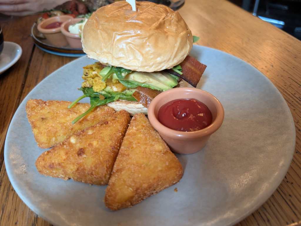 A plate with three crispy hash browns, a pot of sauce, and a burger containing tofu omelette, avocado, and vegan bacon amongst other ingredients