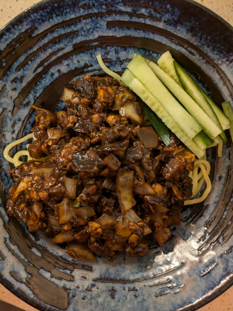 Noodles topped with vegetables and tempeh in a very dark sauce with cucumber sticks