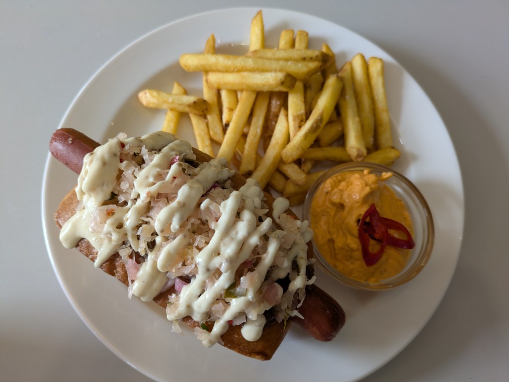 A hotdog with a serving of chips and pot of sauce. The hotdog is piled high with toppings