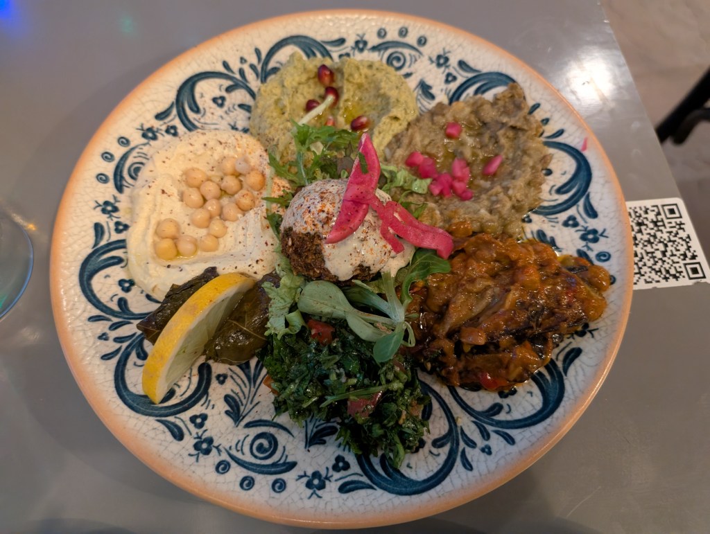 A large plate with one big falafel in the middles, surrounded by two stuffed vine leaves, some tabbouleh, and various dips