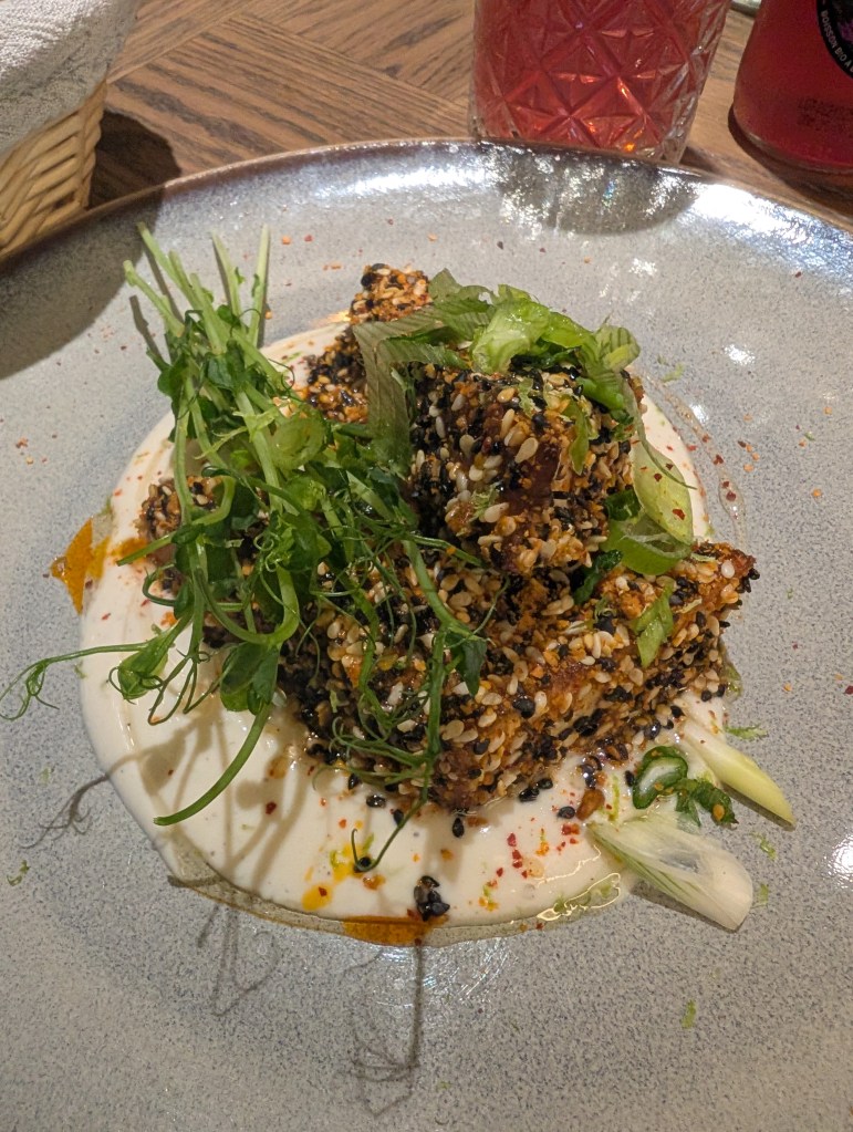 Blocks of tofu crusted in sesame seeds, sitting on a pool of yoghurt and garnished with greens 
