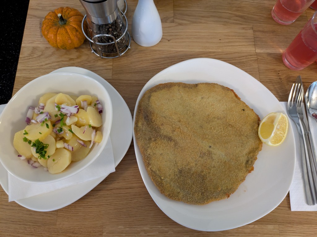 A gigantic schnitzel on a plate with a separate bowl of potato salad on the side