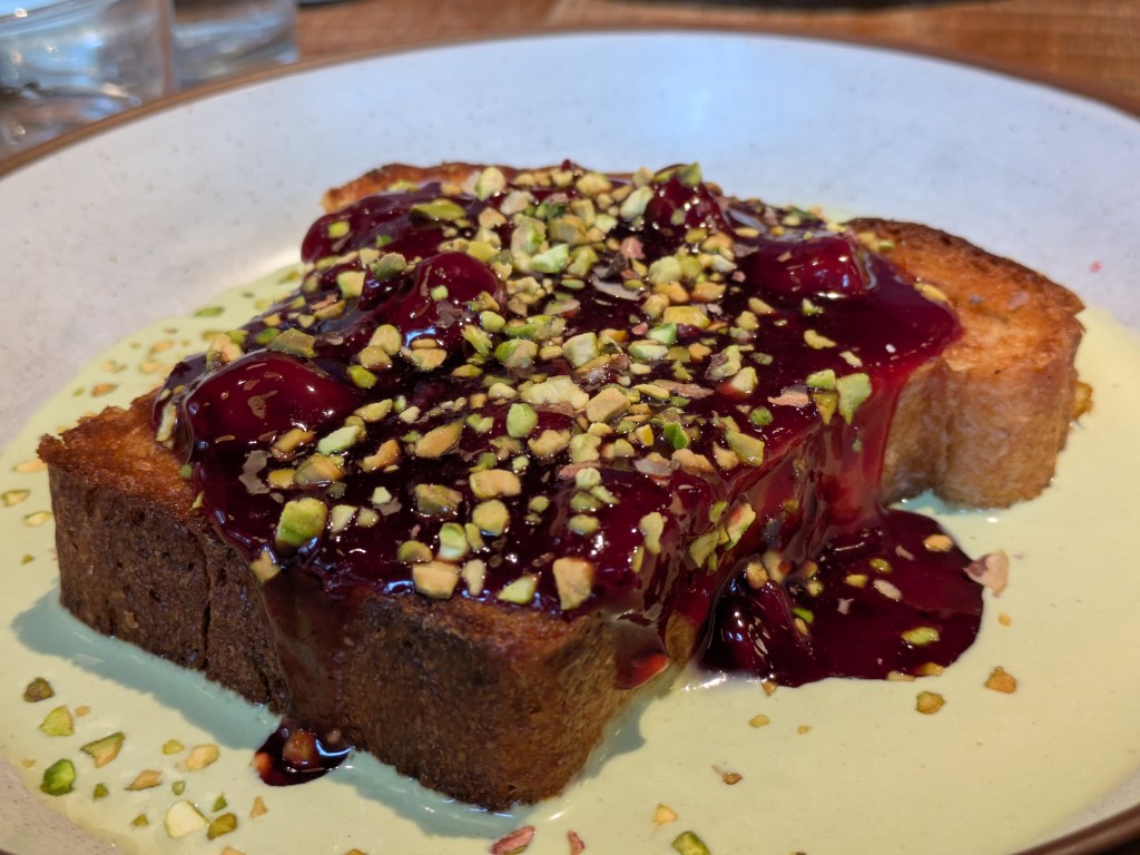 A thick slice of French toast on a pool of pale green custard, topped with a rich jammy red sauce and chopped pistachios 