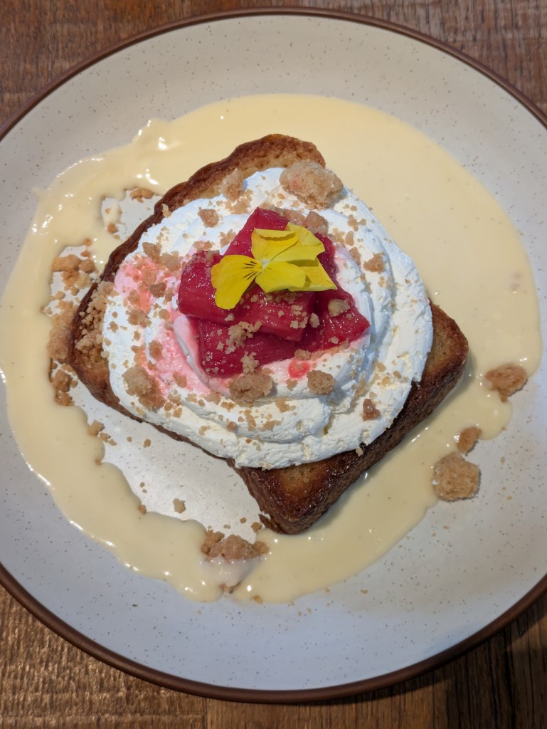 A thick slice of French toast topped with a generous spiral of cream and some pink rhubarb, all sitting in a pool of custard