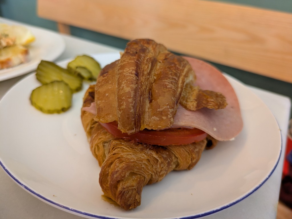 A croissant with a slice each of ham and tomato poking out, with some pickles on the side