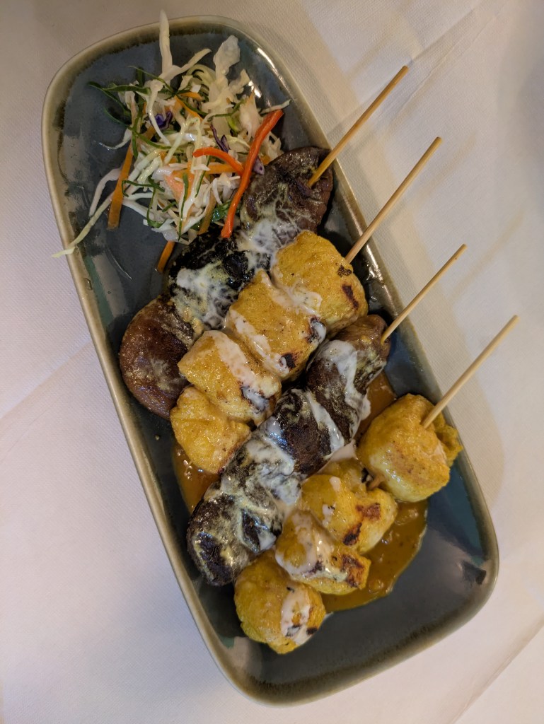 Four skewers, two with chunks of tofu and two with chunks of mushroom, drizzled with sauce alongside a small salad