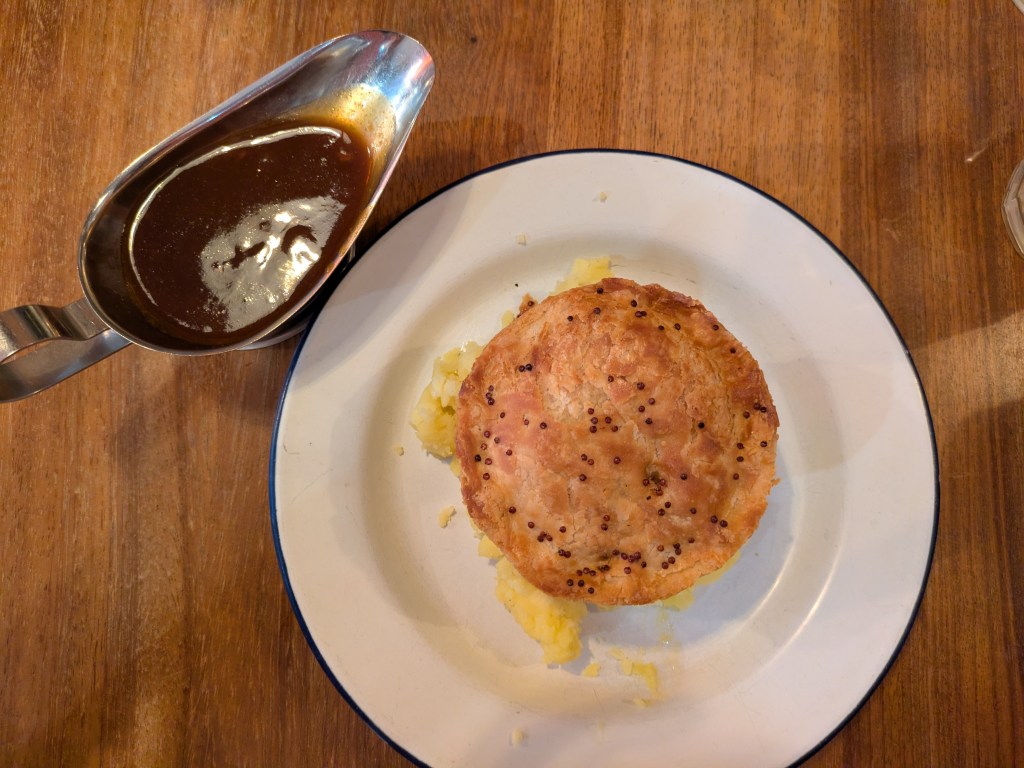 A pie sitting on top of a pile of mashed potato, with a gravy boat on the side 